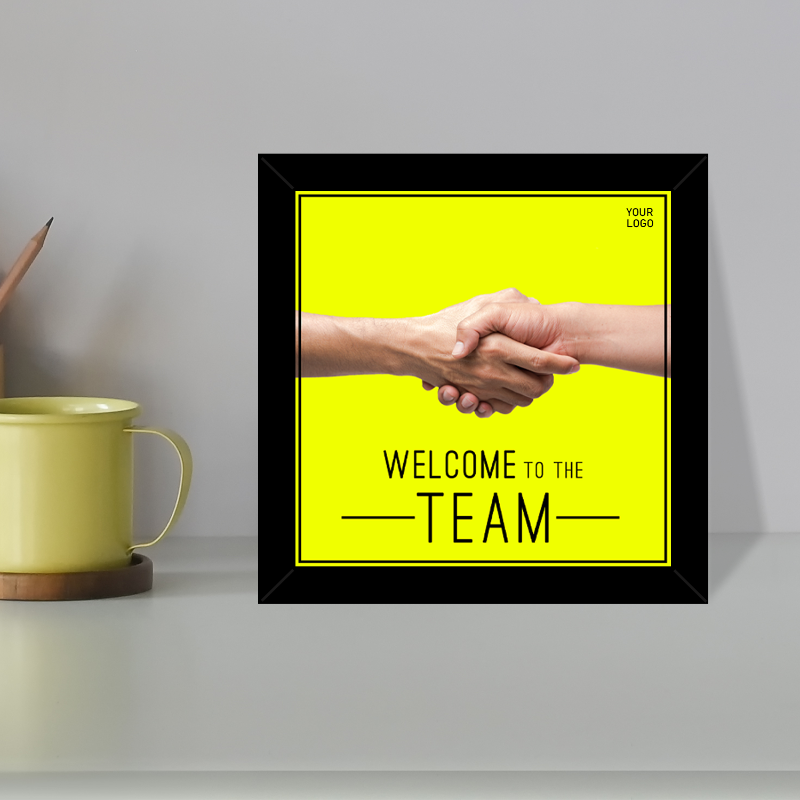 A framed welcome sign with a yellow background and black text that reads 'WELCOME TO THE TEAM', with a handshake graphic. The frame is displayed on a shelf next to a cup and a pen.