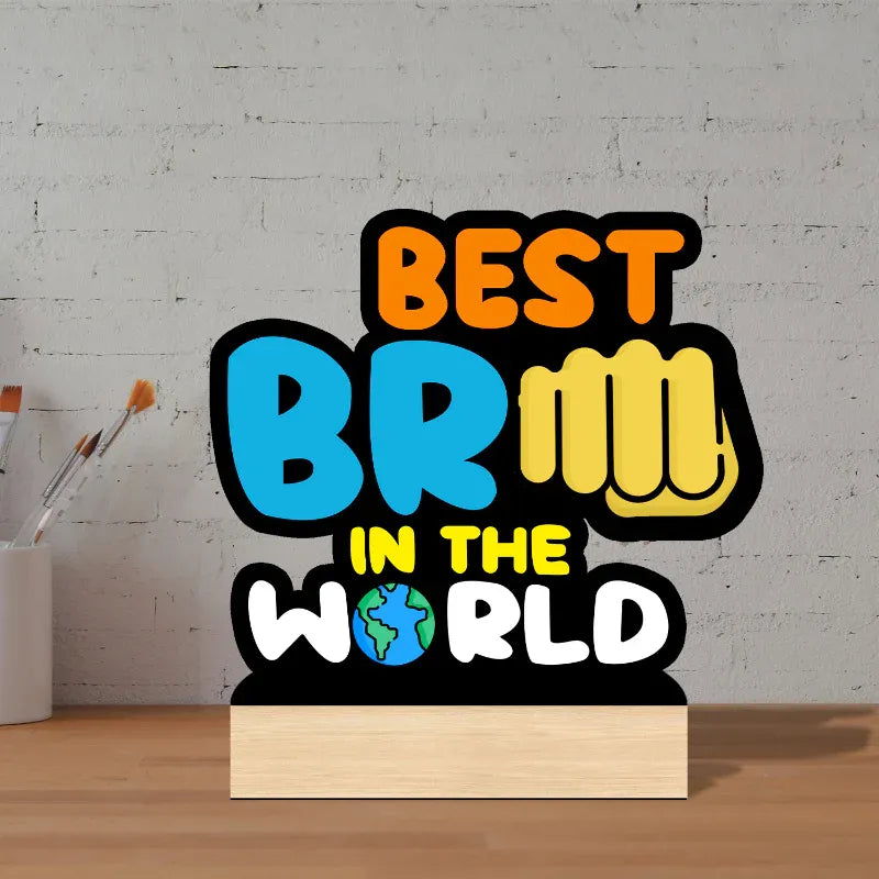 A novelty desk lamp shaped like a book, featuring the text 'BEST BRILLIANT IN THE WORLD' in colorful bubble lettering on a black background, sitting on a light wood surface against a white brick wall.