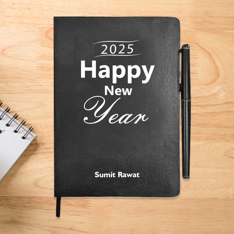 A black journal diary with '2025 Happy New Year' text on the cover, alongside a pen, on a wooden surface. There is also a spiral notebook to the left.
