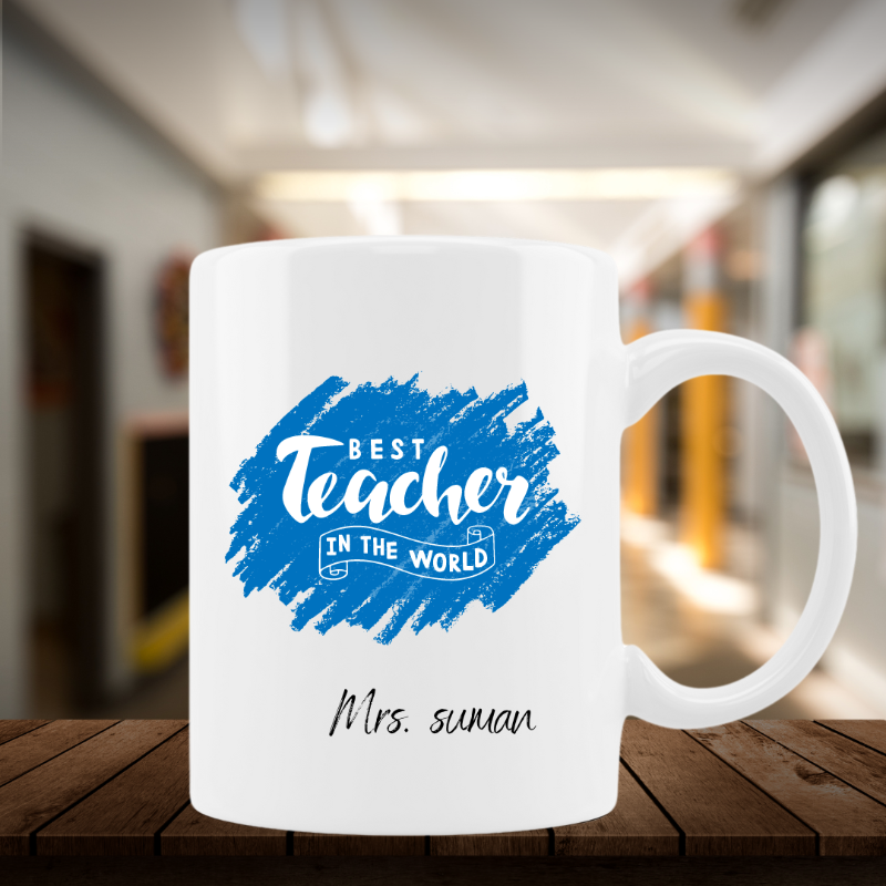 A white ceramic mug with a blue and multicolor design that reads 'Best Teacher in the World', with a name 'Mrs Suman' printed on it, placed on a wooden surface with a blurred background.
