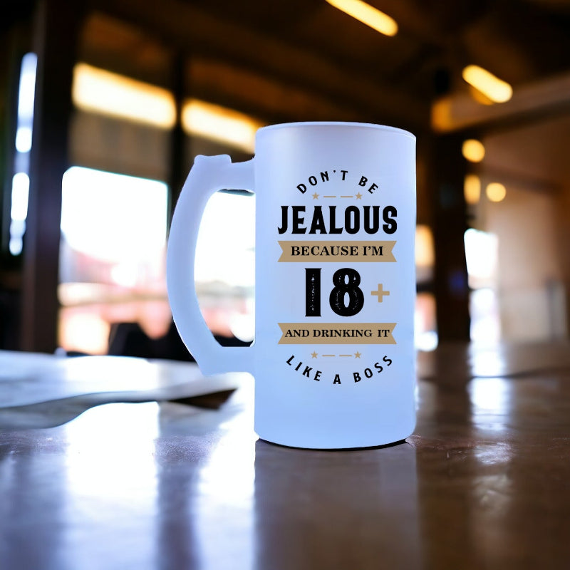 A white beer mug with the text 'DON'T BE JEALOUS BECAUSE I'M 18+ AND DRINKING IT' printed on it, placed on a wooden surface.