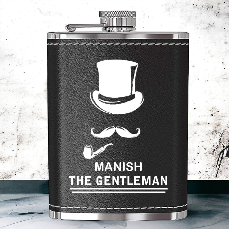 A black leather hip flask with a stainless steel cap, personalized engraving that reads 'MANISH THE GENTLEMAN' along with an illustration of a top hat and a mustache.
