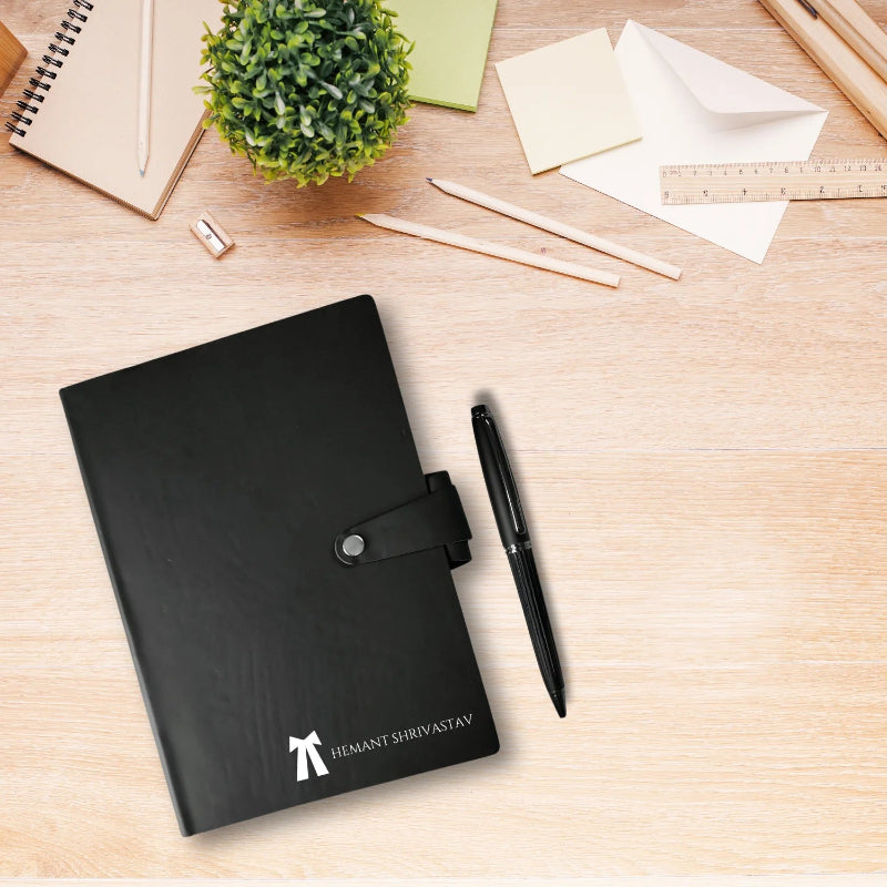 A personalized black diary with a pen, placed on a desk surrounded by stationery items, indicating the product is a diary with a sleek black leatherette cover and a pen included.