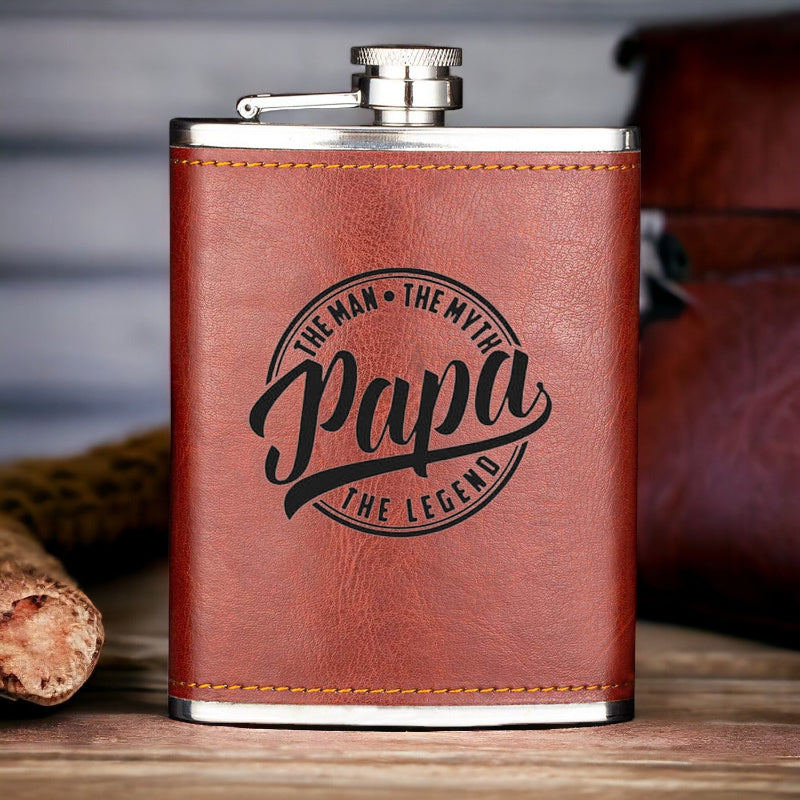 A tan leather hip flask with 'PAPA THE LEGEND' printed in black text, placed on a wooden surface next to a brown leather bag.