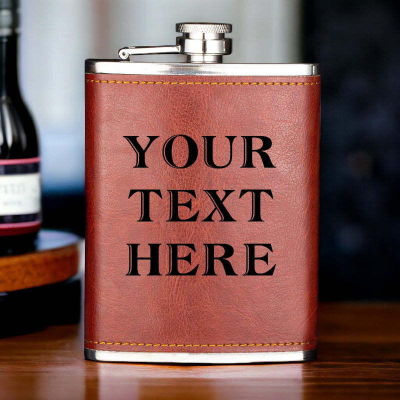 A tan leather hip flask with personalized engraving, resting on a wooden surface, with a blurred background suggesting a celebratory setting.