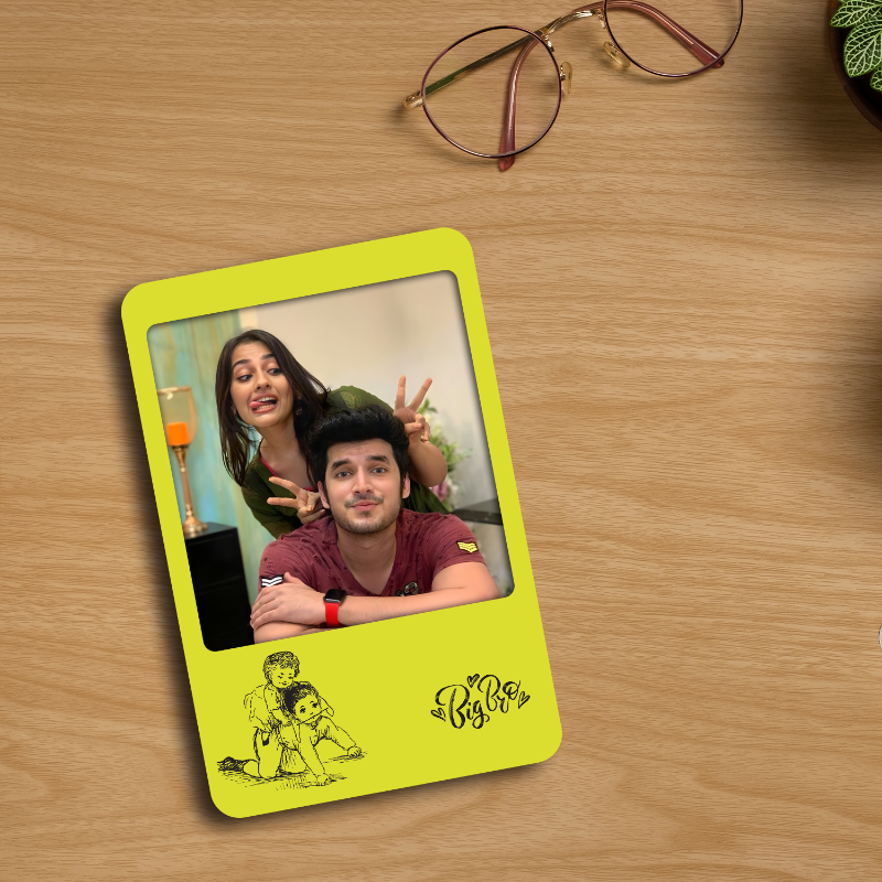 A personalized yellow fridge magnet with a photo of a brother and sister, along with text, placed on a wooden surface next to glasses and a plant.