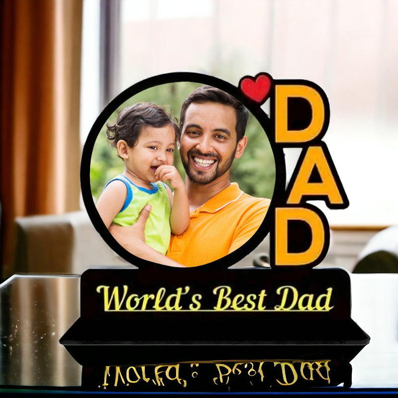 A personalized picture frame with the inscription 'World's Best Dad' at the bottom, featuring an image of a smiling adult with a child. The frame appears to be made of black metal or acrylic.