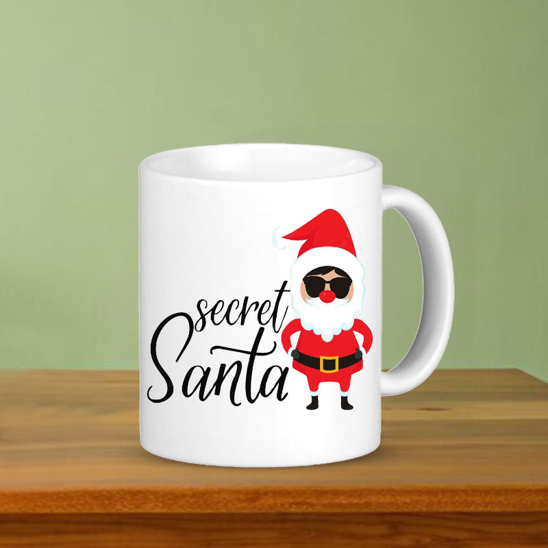 White mug with a cartoon Santa Claus design and 'secret Santa' text on a wooden surface with green background
