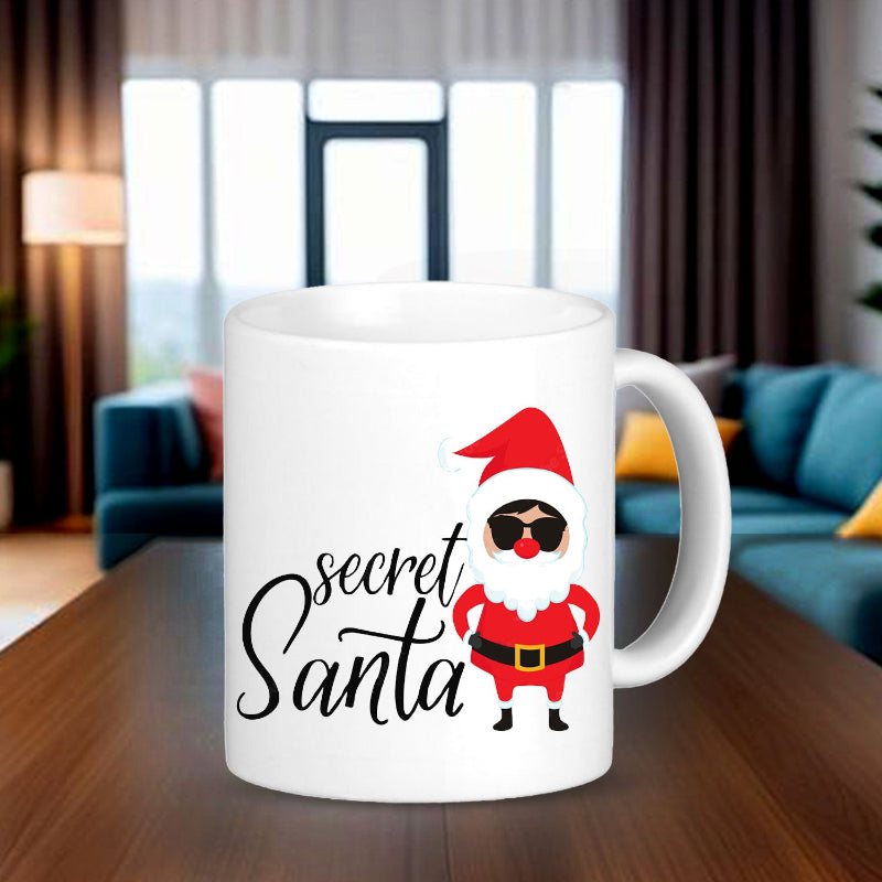 A white ceramic mug with a printed design featuring the text 'Secret Santa' and an illustration of Santa Claus wearing sunglasses, placed on a table with a cozy room and blurred Christmas decoration in the background.
