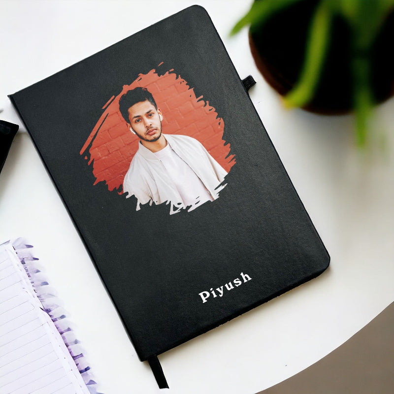 Black notebook with a portrait of a man on the cover and 'Piyush' written below, placed on a white surface.