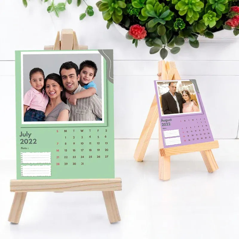 Two desk calendars with family photos on wooden stands, one green and one purple, with a plant in the background.