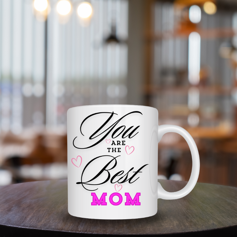 White mug with 'You are the Best Mom' text on a wooden table in a blurred cafe setting