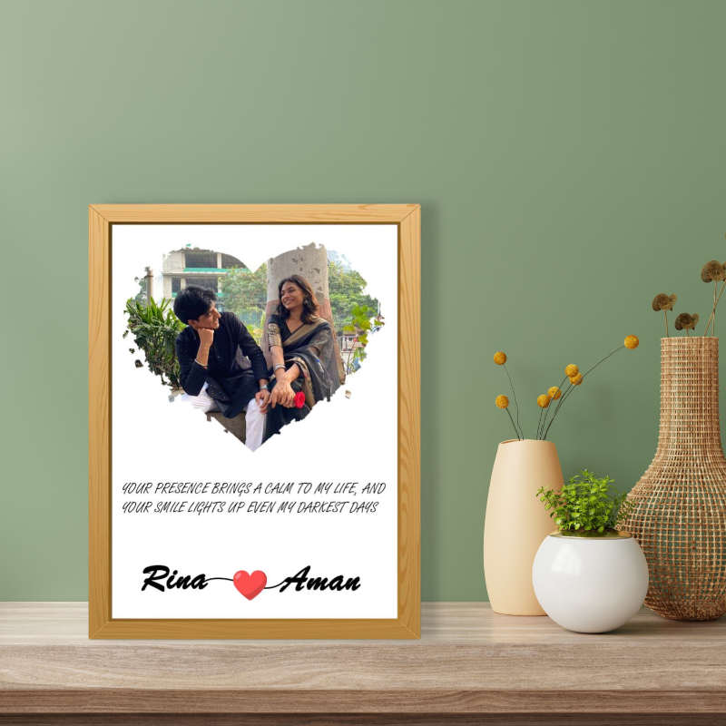 An elegant wooden picture frame with a heart-shaped photo window, displaying a couple's picture. The frame is placed on a wooden table with a green background, alongside a vase with dried flowers.