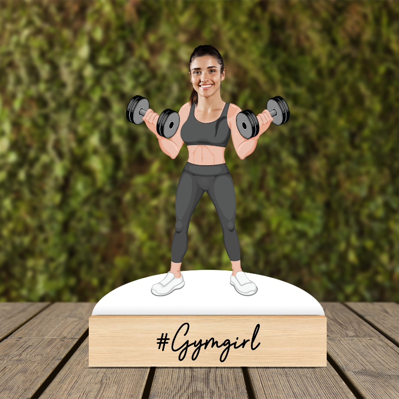 Figurine of a woman lifting weights with '#Gymgirl' text on a wooden base against a green foliage background.