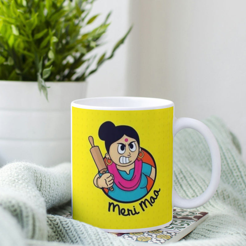 Yellow mug with a cartoon character and 'Meri Maa' text, placed on a surface with a plant in the background.