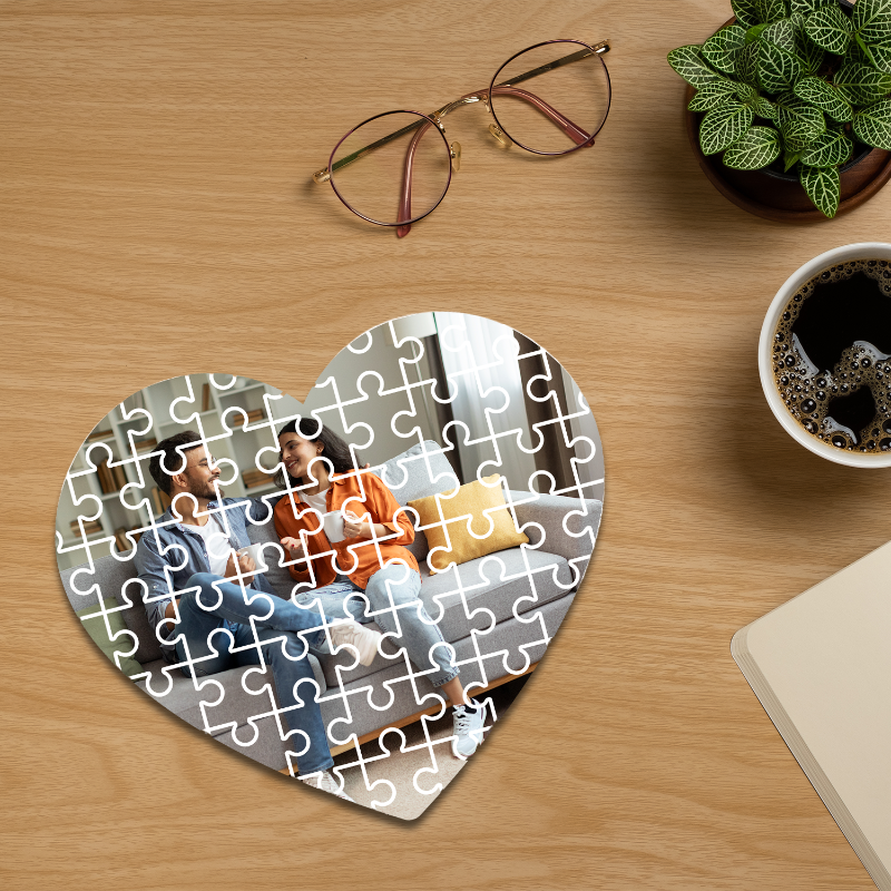 Heart-shaped puzzle on a wooden surface with glasses, plant, and coffee cup in the background
