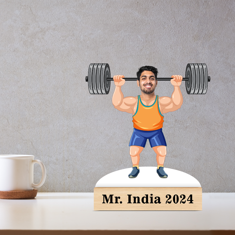 Figurine of a man lifting weights with 'Mr. India 2024' on a base, placed on a surface with a mug in the background.