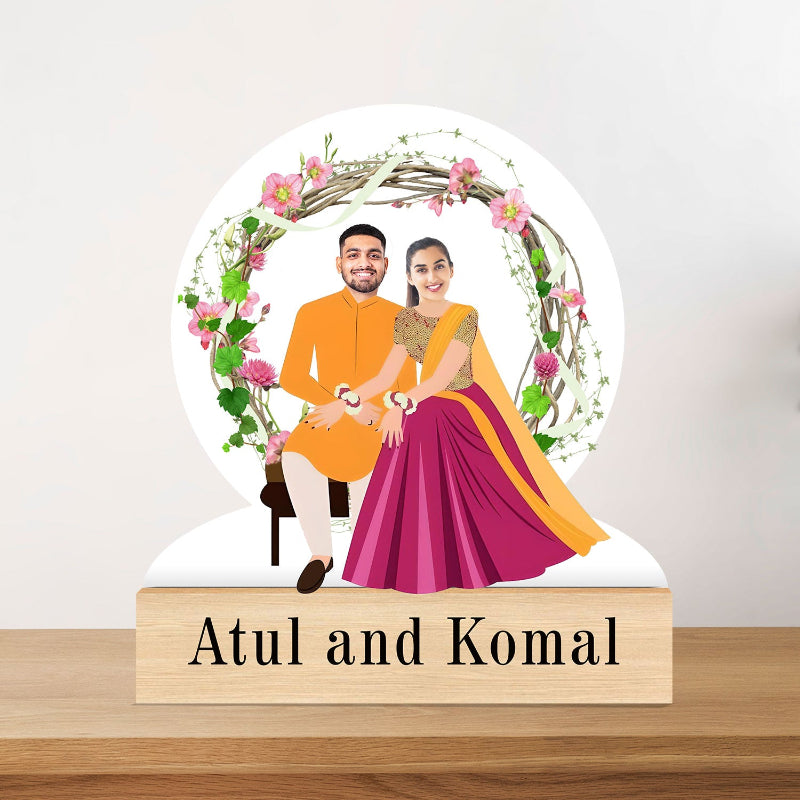 Decorative display with a couple in traditional attire surrounded by floral elements, on a wooden base with 'Atul and Komal' text.