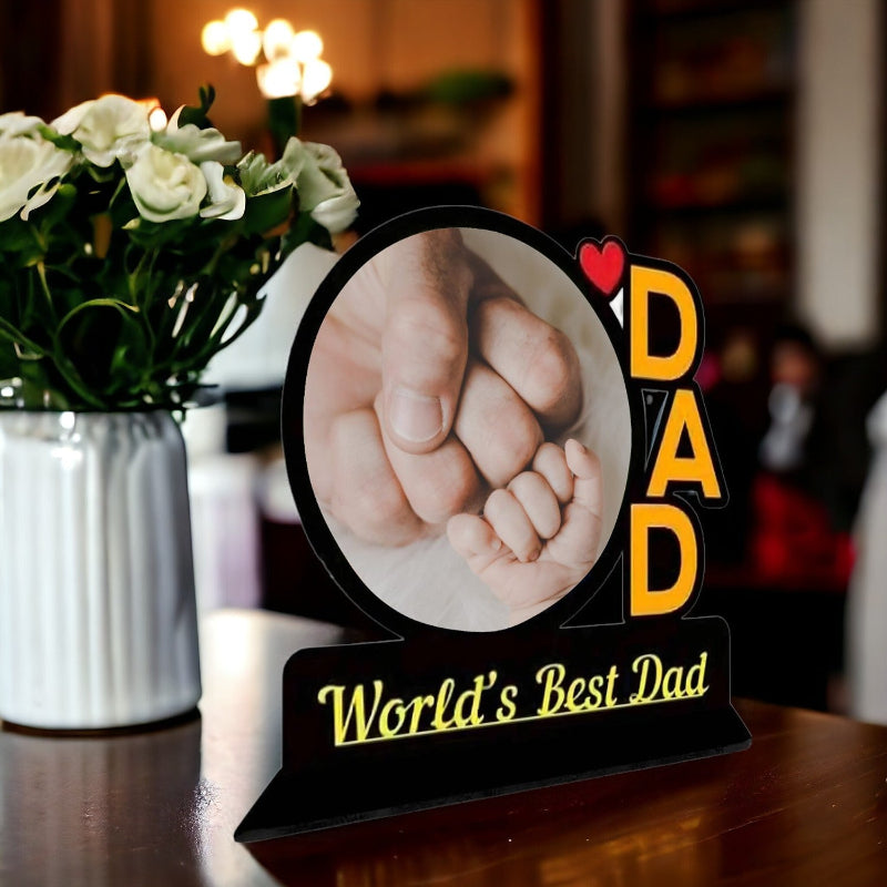 Decorative sign with 'DAD' and 'World's Best Dad' text, featuring a photo of two fists holding hands, on a blurred background with flowers.
