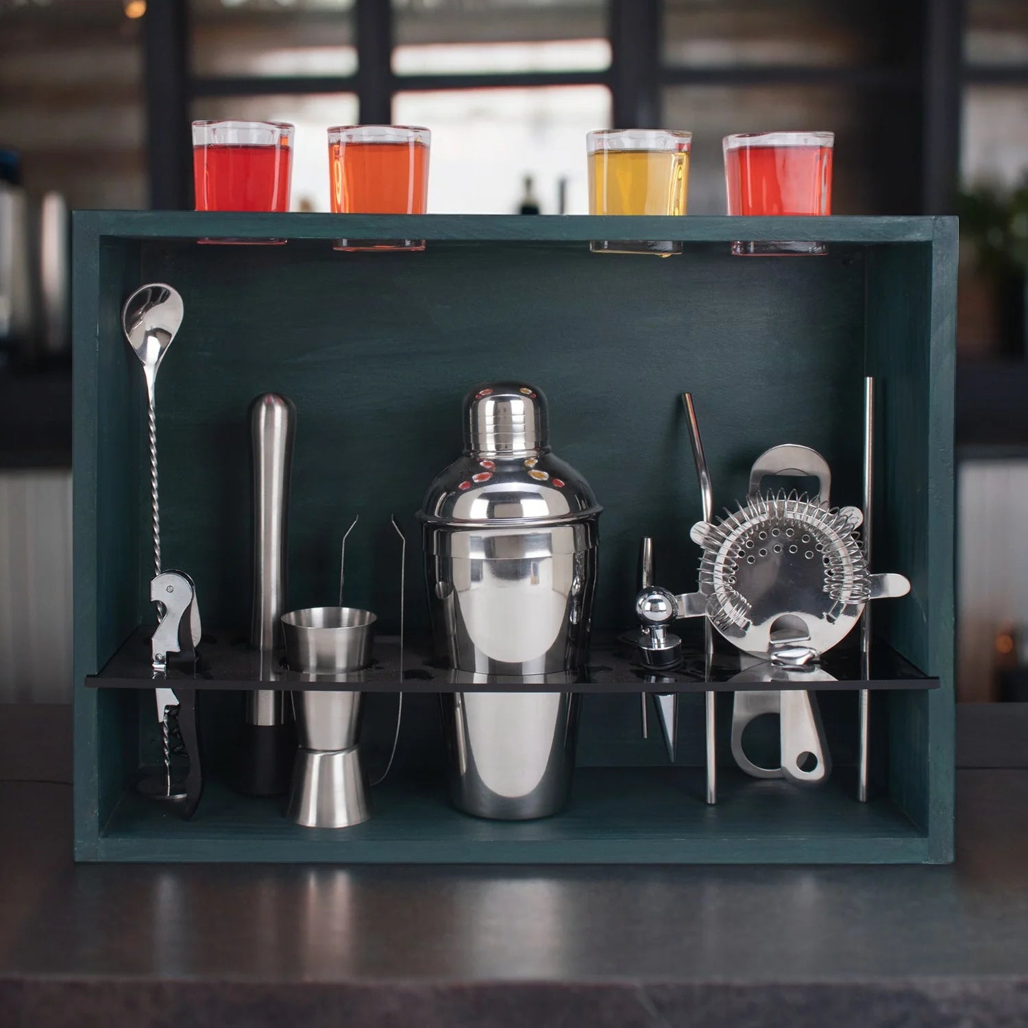 A 17-piece home bar kit displayed on a military grey wall stand, including cocktail shaker, muddler, jigger, mixing spoons, strainers, pourers, tongs, bottle opener, corkscrew, zester/peeler, wine stopper, and four shot glasses.