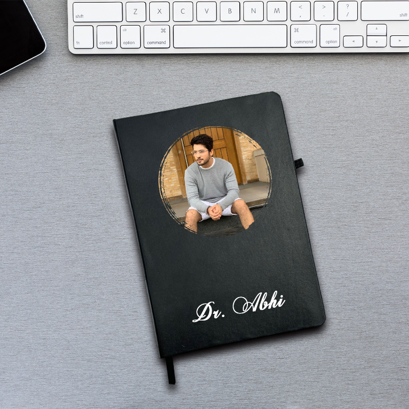 Black notebook with a photo of a man on the cover, placed on a desk with a keyboard.