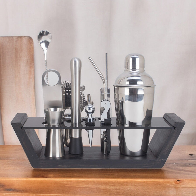 A 12-piece bar set displayed on a sleek wooden stand, including a cocktail shaker, muddler, jigger, mixing spoon, strainer, pourers, tongs, bottle opener, cork screw, two straws, and a wine stopper, all arranged neatly on a wooden table.