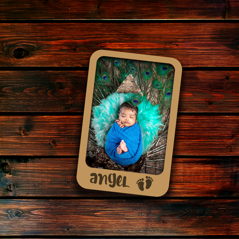 Newborn baby in a blue outfit surrounded by peacock feathers with 'angel' text on a wooden background