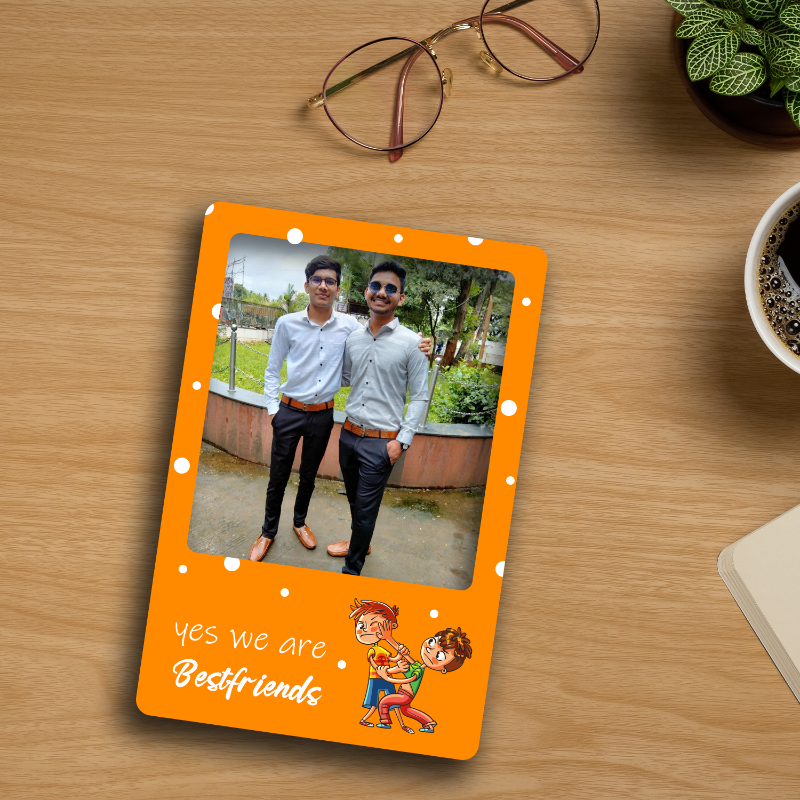 A personalized orange fridge magnet with a photo of two men and text on a wooden surface. The magnet has a 3D effect with a white background and is placed on a wooden refrigerator.