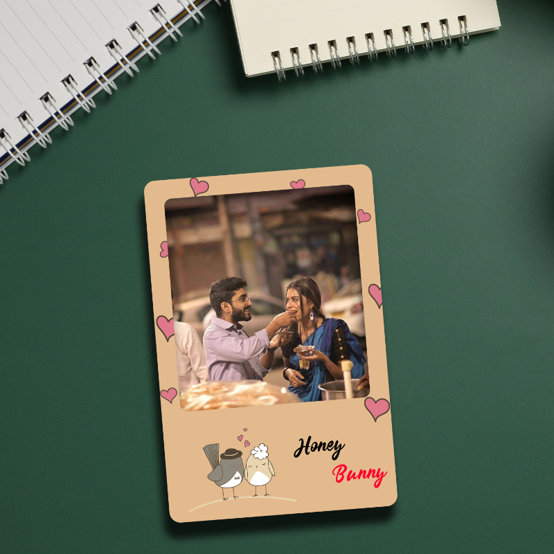 A personalized acrylic fridge magnet with a photo of a couple and decorative elements such as hearts and a cartoon bunny, placed on a green surface next to a notebook and a pen.