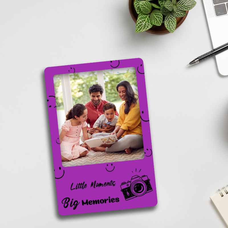 A purple acrylic fridge magnet with a personalized photo of a family on it, placed on a refrigerator with a laptop, notebook, and potted plant in the background.