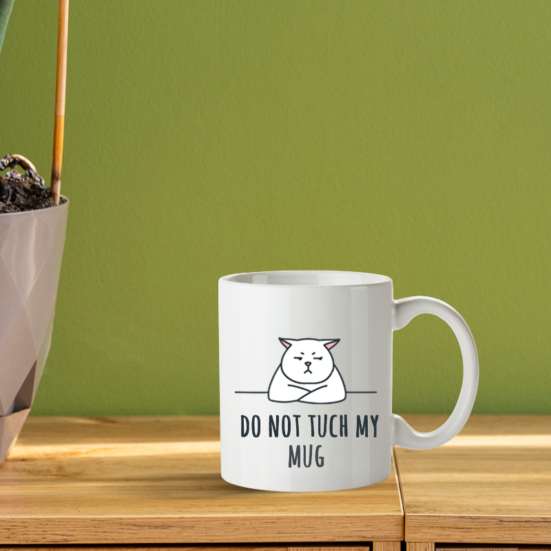 White mug with a cat illustration and text on a wooden surface with a green wall background