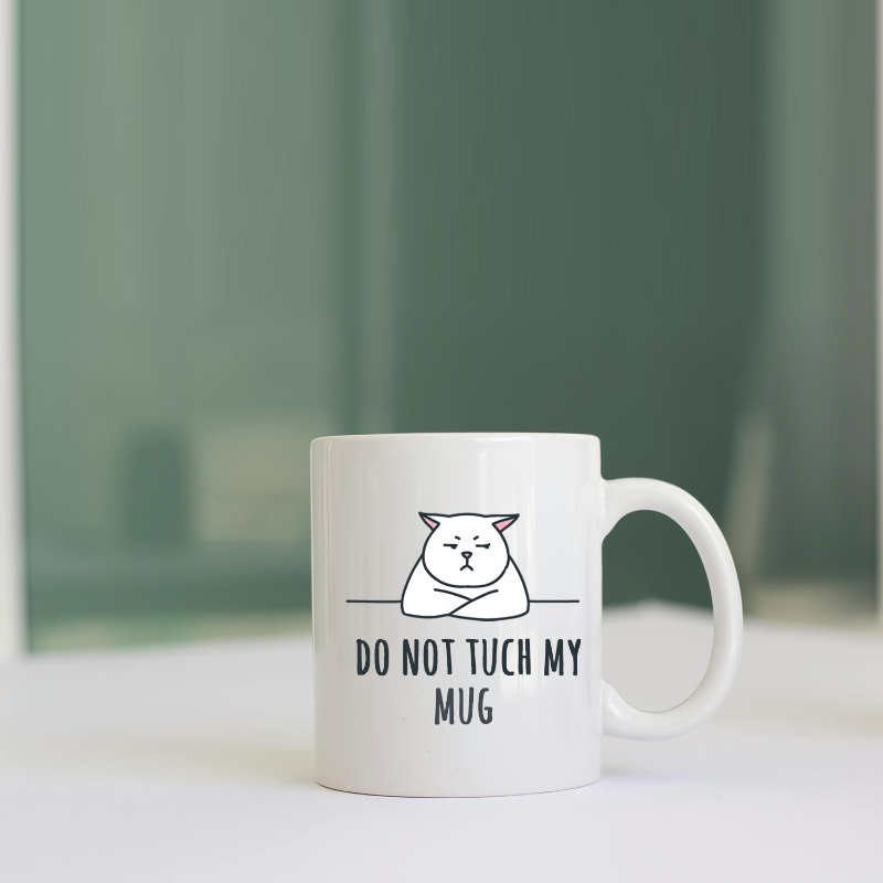 White mug with a cat illustration and text 'Do Not Touch My Mug' on a white surface with a green blurred background.
