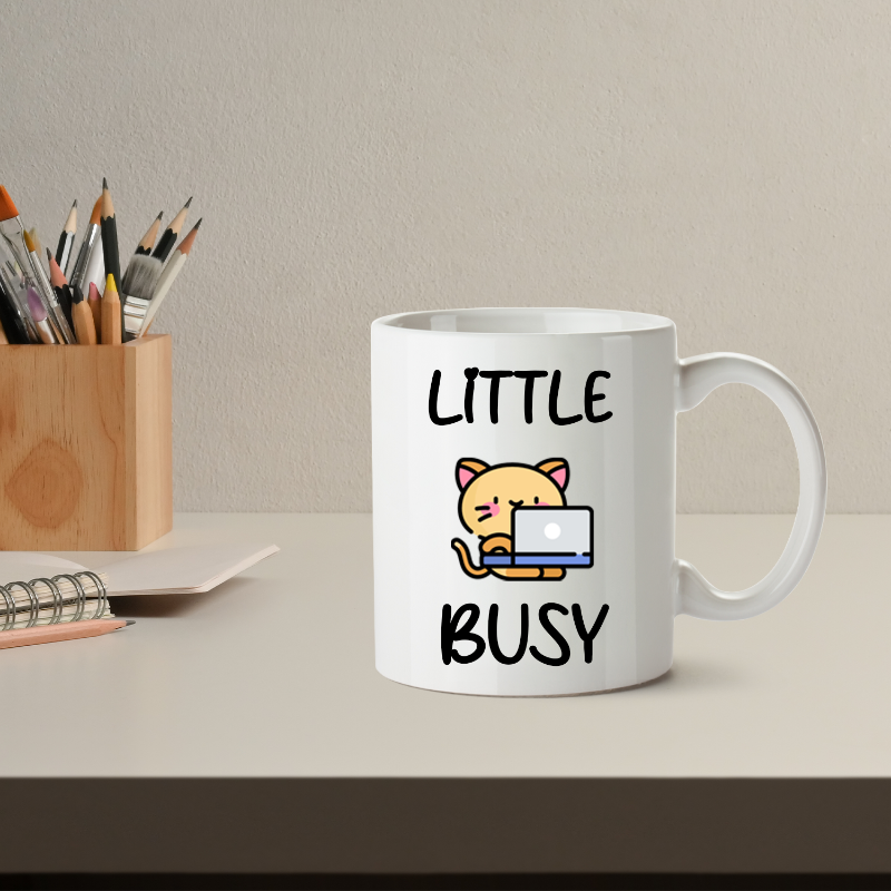 White mug with a cat illustration and text 'LITTLE BUSY' on a desk with office supplies.