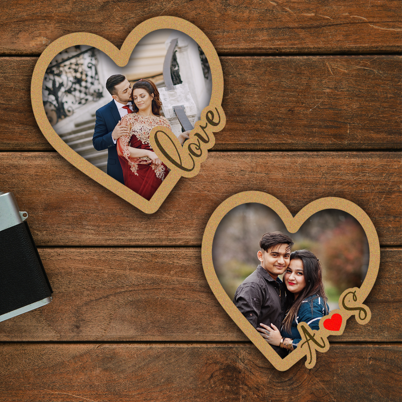 Two personalized heart-shaped couple photo fridge magnets on a wooden surface. One magnet has a photo of a couple and the word 'Love' written on it, while the other has a photo of a man and a woman holding hands with the word 'AS' written on it.
