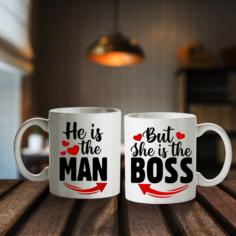 Two white ceramic mugs with printed text, one with the text 'HE IS THE MAN' and the other with 'BUT SHE IS THE BOSS', placed on a wooden surface with a blurred indoor background.