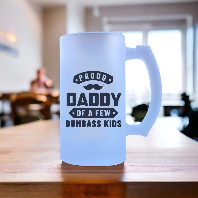 A white ceramic beer mug with a printed phrase 'PROUD DADDY OF A FEW DUMBASS KIDS' on it, placed on a wooden table with a blurred indoor background.
