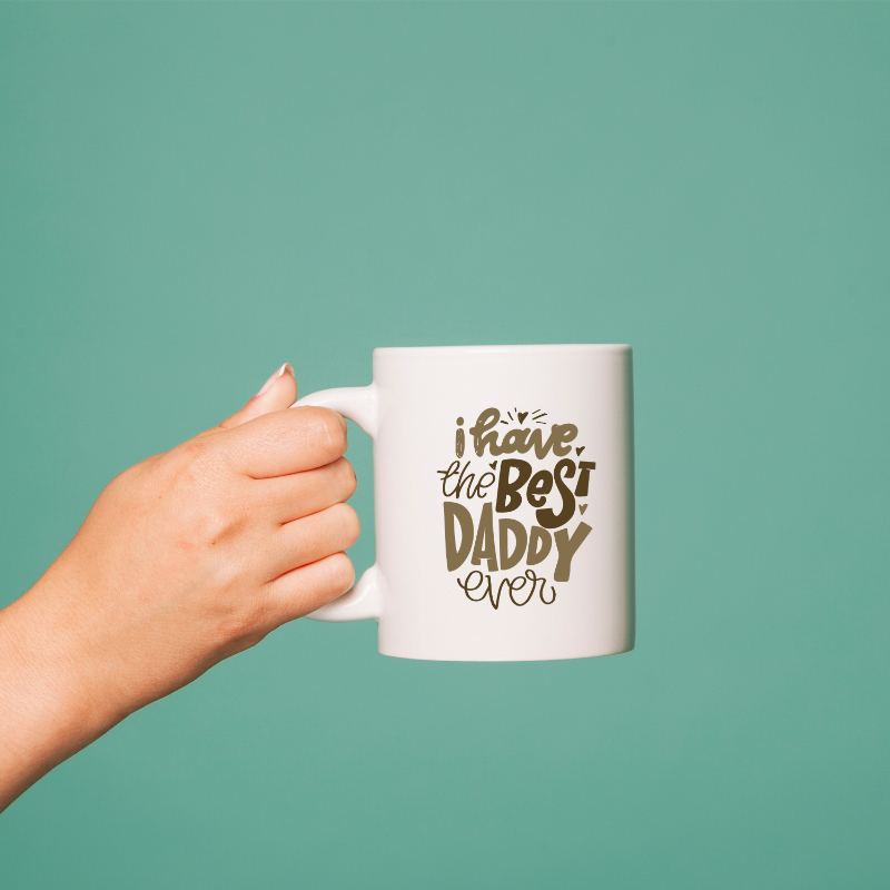 A hand holding a white ceramic mug with text printed on it, against a teal background.