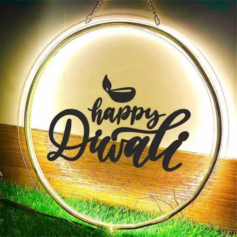 A glowing round Diwali LED Light sign with a gold-toned frame and black script reading 'happy Diwali' on a light background, displayed outdoors on artificial grass against a dark backdrop.