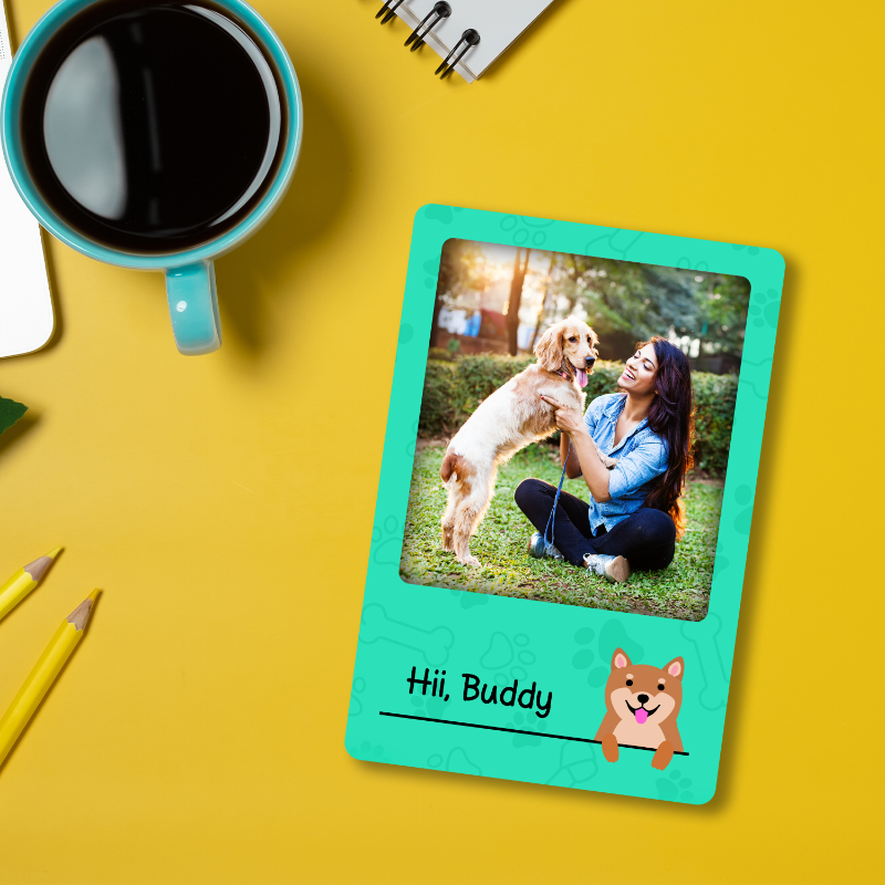 A customized fridge magnet with a photo of a person and a dog, placed on a yellow surface next to a blue cup and yellow pencils.