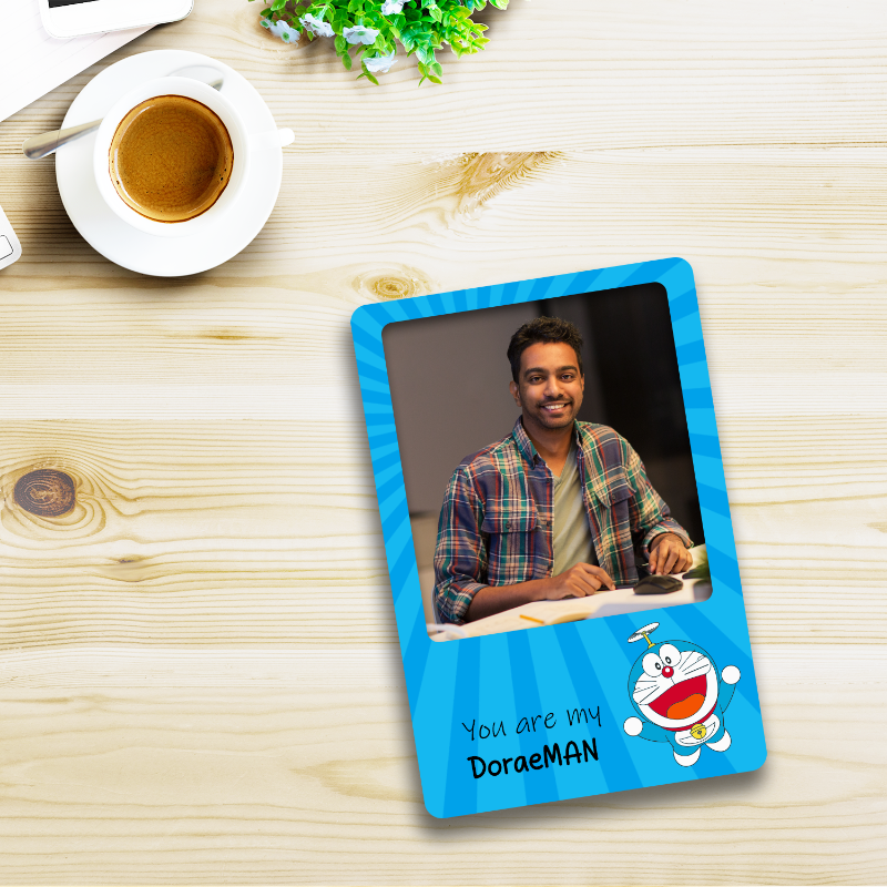 A blue acrylic fridge magnet with a personal photo and text on it, placed on a wooden surface with a cup of coffee and decorative items around.