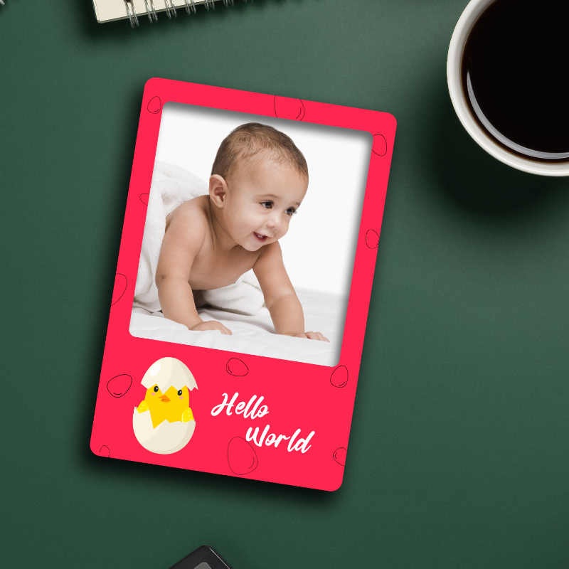 A personalized acrylic fridge magnet with a baby's photo and the text 'Hello World' on a green background, with a cup of coffee and a notebook in the background.