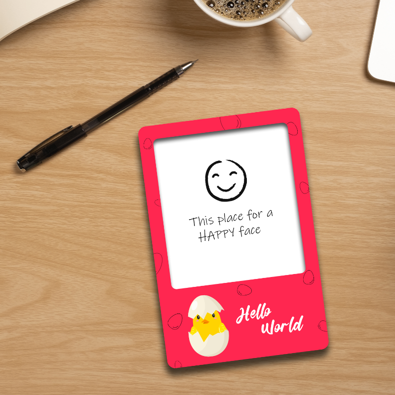 Small digital display with a smiley face and text on a wooden surface with a pen and coffee cup.
