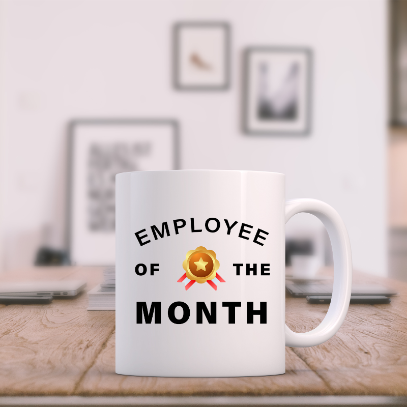 A white ceramic mug with the text 'EMPLOYEE OF THE MONTH' and a gold star emblem on a wooden table in an office setting.