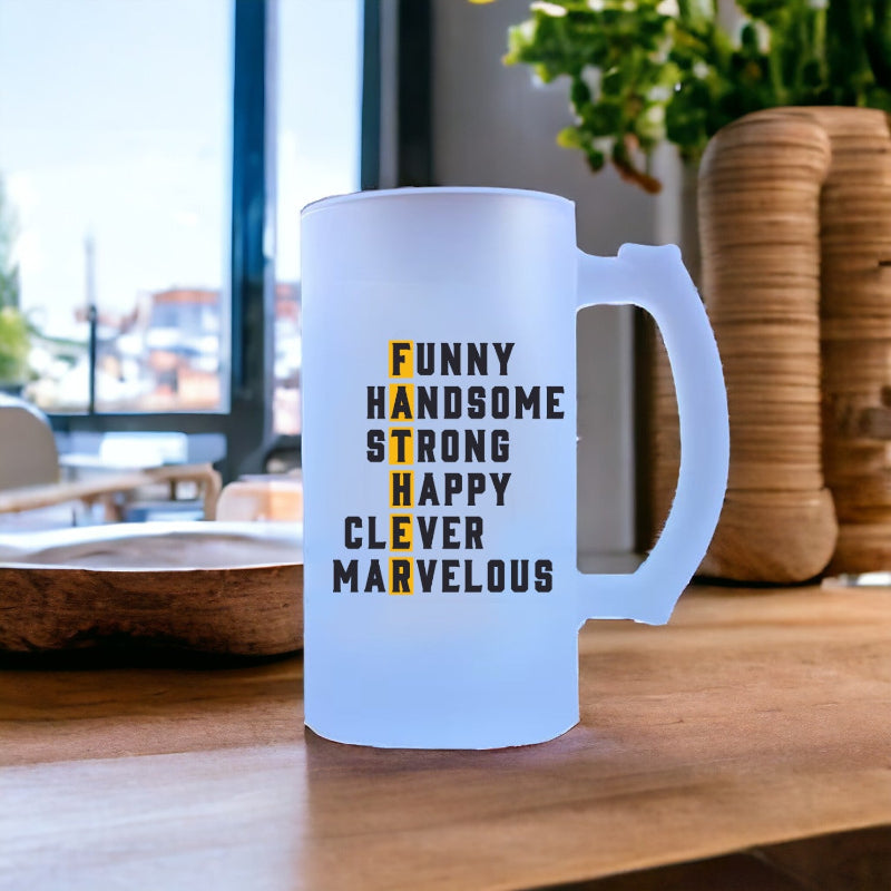 A white premium motifc print beer mug with black and yellow text, placed on a table with a plant and a wooden object in the background, indicating the text on the mug which says 'Funny, Handsome, Strong, Happy, Clever, Marvelous'.