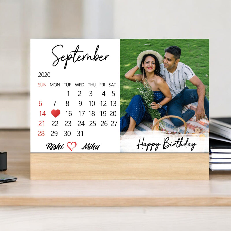 A personalized birthday calendar card with a photo of a couple, standing on a desk with a wooden background. The card has a 'September 2020' header, and each day is marked with a number from 1 to 30, along with 'Happy Birthday' text and the names 'Rahul' and 'Nikhil'.