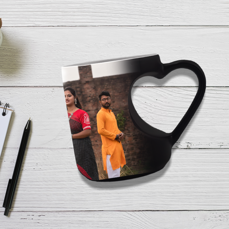 A heart-shaped ceramic mug on a white wooden surface with a photograph of two people, possibly a couple, in the background. The mug has a black exterior and reveals a photo when filled with a hot beverage.