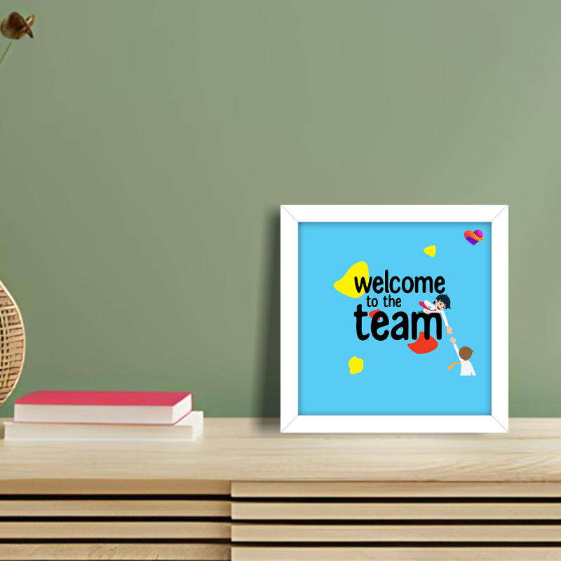 Framed artwork with 'Welcome to the team' text on a wooden shelf against a green wall.