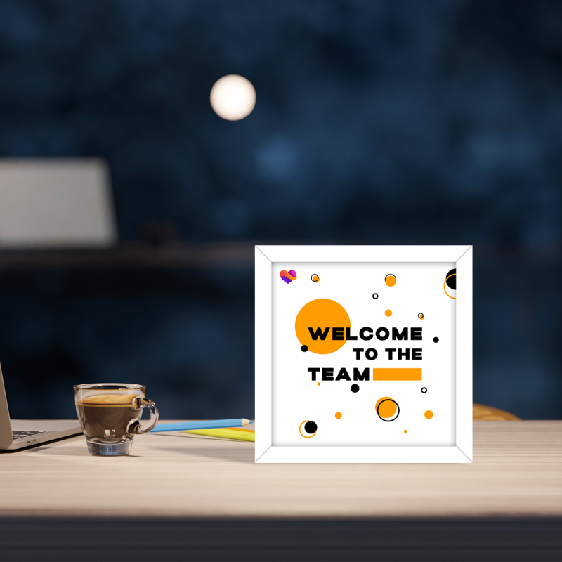 Framed 'Welcome to the Team' sign on a desk with a laptop and coffee cup in the background.