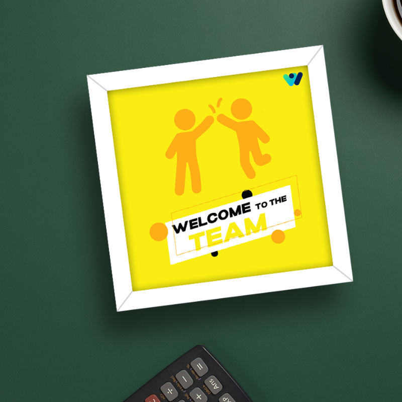 A yellow rectangular frame with the text 'Welcome to the TEAM' and an illustration of two hands high-fiving each other on the cover, placed on a green surface with a remote and a cup in the background.