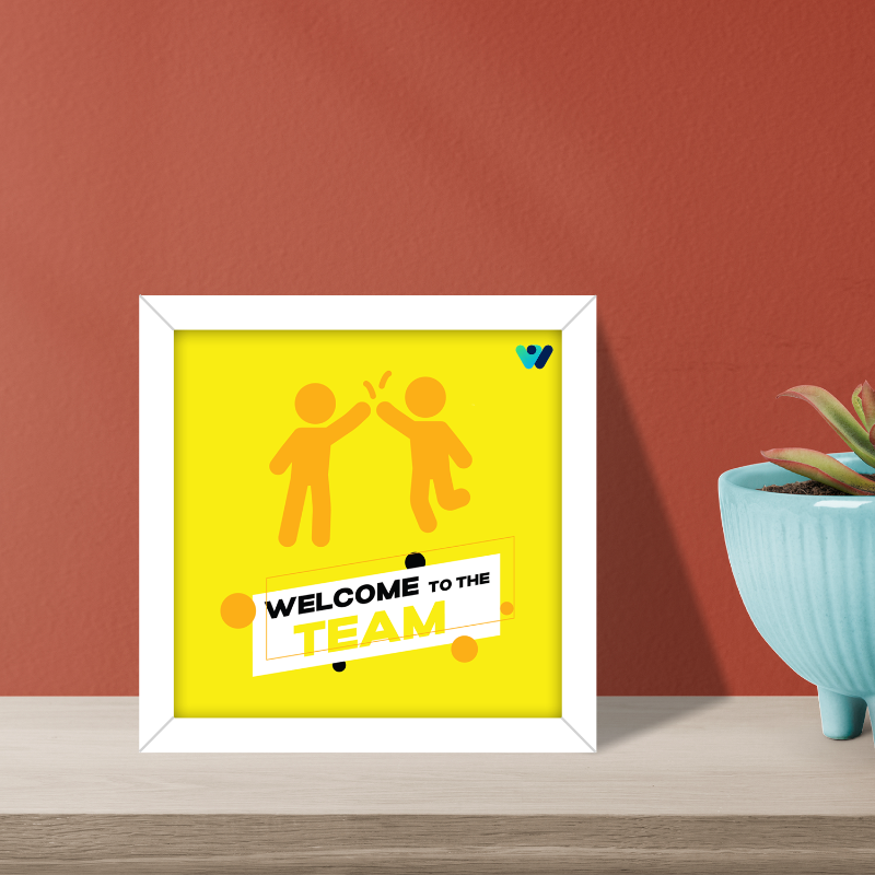Framed poster with 'Welcome to the Team' text and two people giving a high-five on a yellow background, placed on a wooden surface with a red wall and plant in the background.